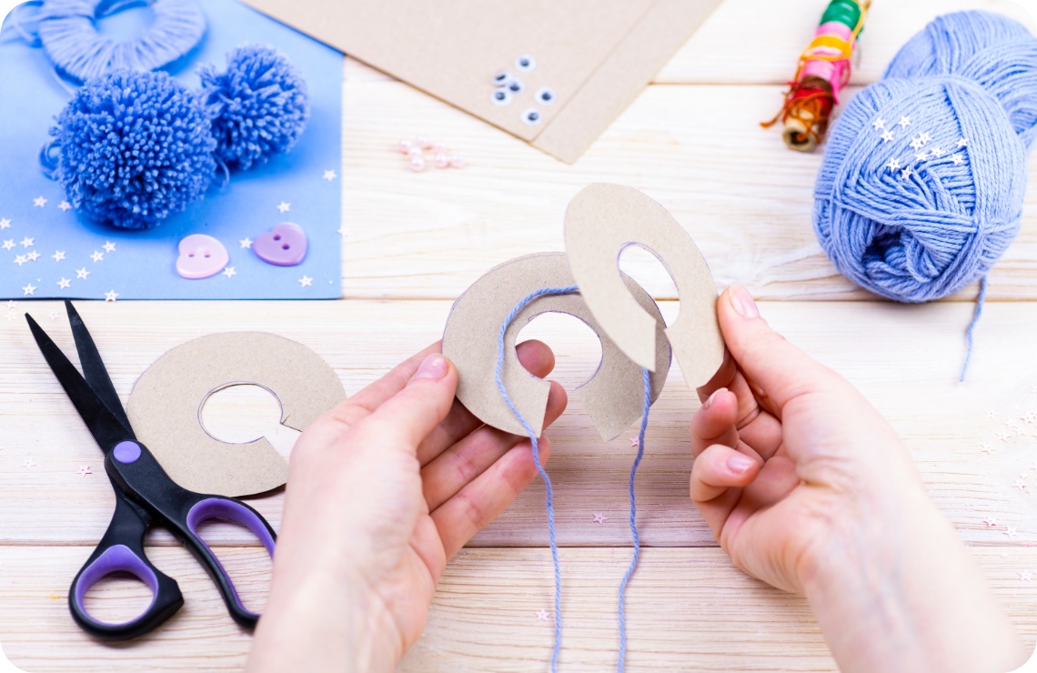 Hände wickeln blaue Wolle um Pappschablonen zur Herstellung von Pompons. Bastelmaterialien auf hellem Holztisch.