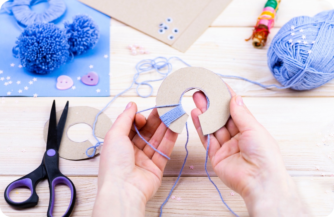 Hände wickeln blaue Wolle um eine Pappschablone für Pompons. Bastelmaterialien wie Schere und Knöpfe auf Holztisch.