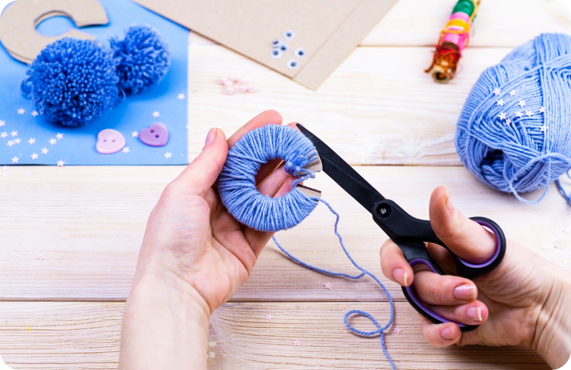 Hände schneiden blaue Wolle auf einer Pompon-Schablone. Bastelmaterialien wie Garn und Schere auf hellem Holztisch.