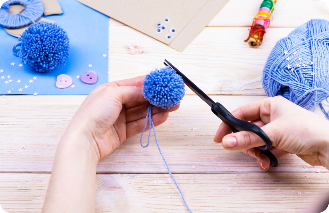 Hände schneiden mit einer Schere einen blauen Woll-Pompon zurecht. Bastelmaterialien auf hellem Holztisch.