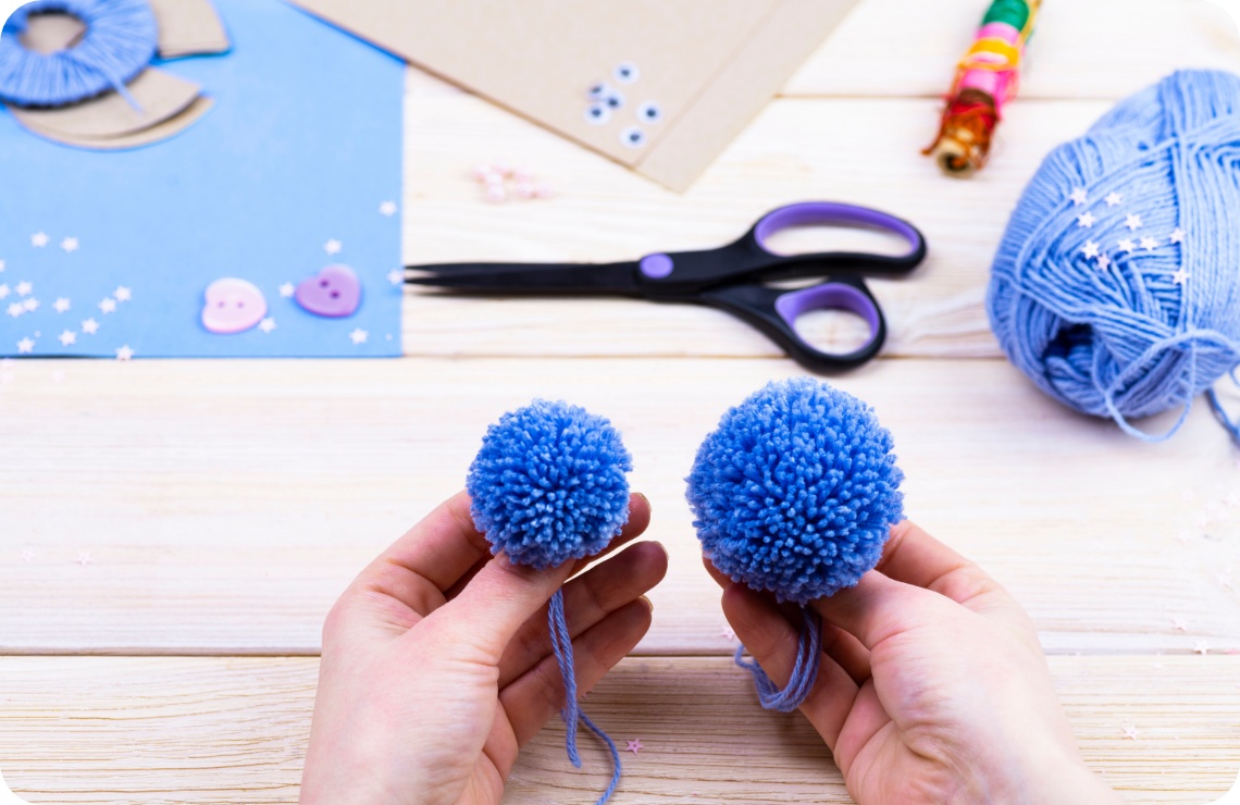 Hände halten zwei unterschiedlich große blaue Woll-Pompons. Bastelmaterialien wie Schere und Garn liegen auf einem Holztisch.