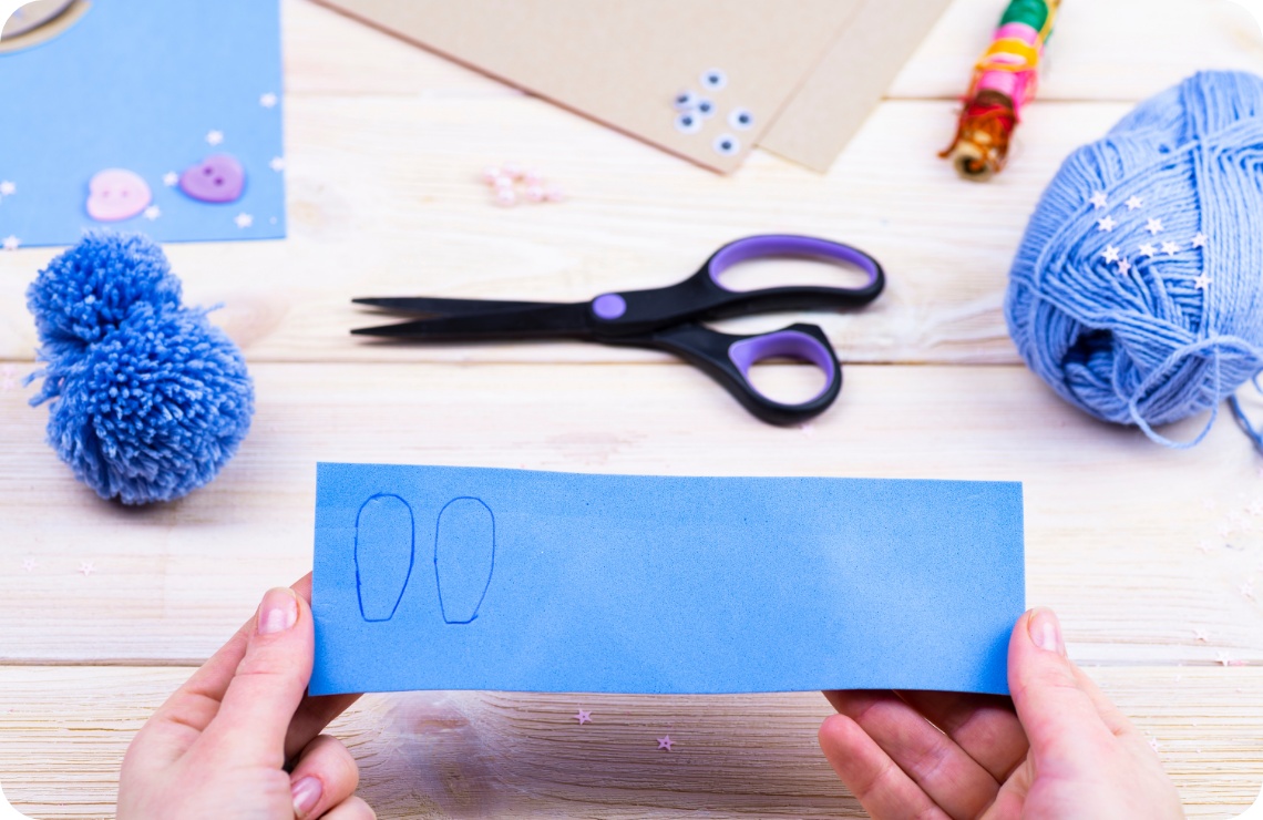 Hände halten blaues Bastelpapier mit aufgezeichneten Hasenohren. Bastelmaterialien wie Schere, Wolle und Pompon liegen auf Holztisch..