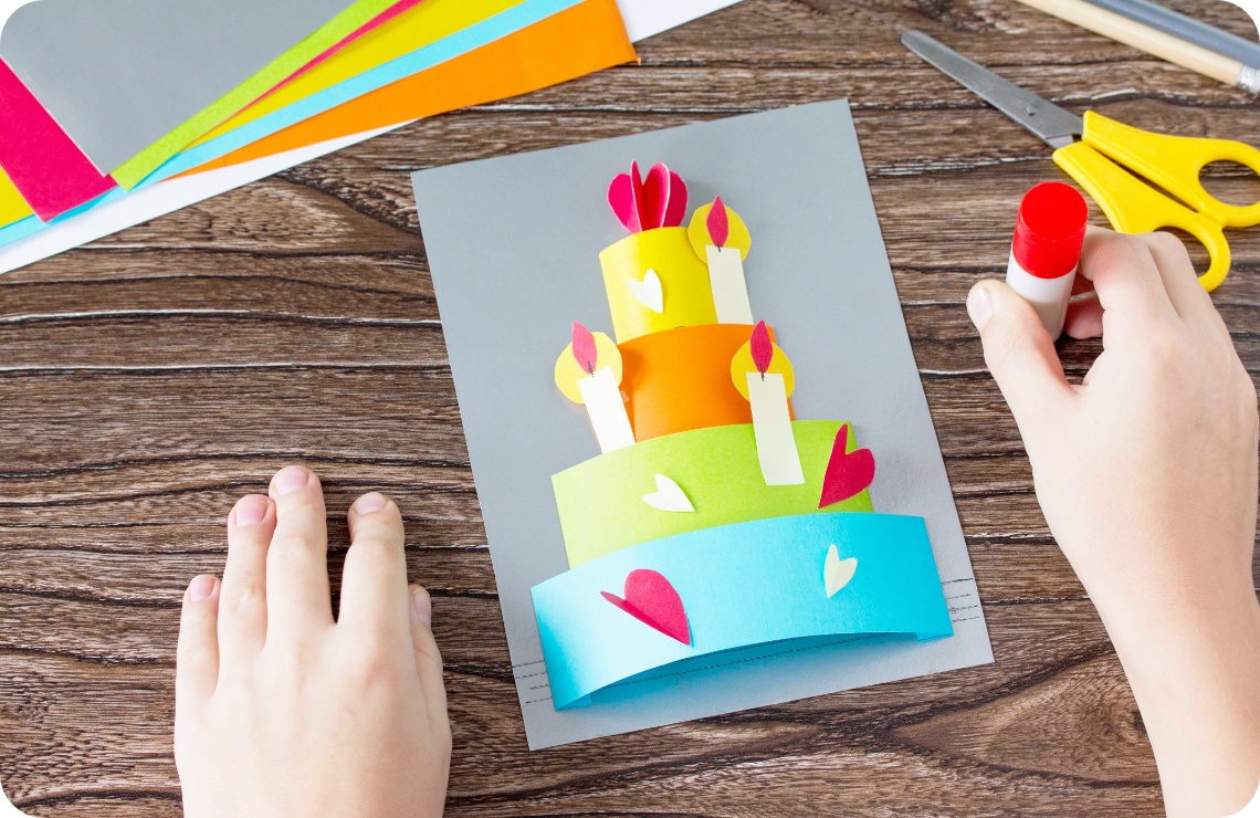 Kinderhände basteln eine bunte 3D-Geburtstagskarte mit einer Papier-Torte und Kerzen. Bastelmaterialien auf Holztisch.