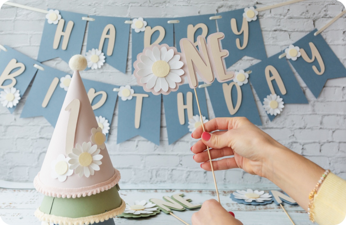 Hand hält eine DIY-Geburtstagsdekoration. Im Hintergrund blaue Happy-Birthday-Girlande und Partyhut.