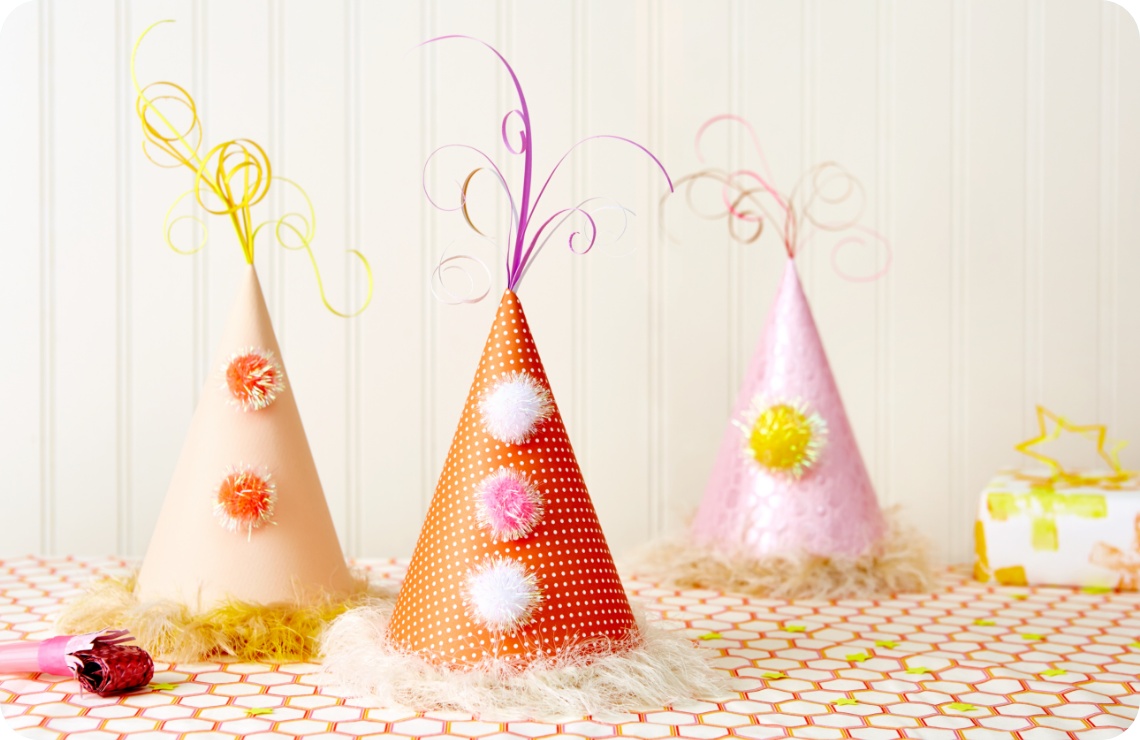 Drei festliche Partyhüte mit Pompons und Lockendekor auf dekoriertem Tisch. Farbenfrohe Geburtstagsdekoration im Hintergrund.