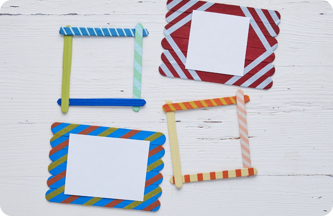 Bunte DIY-Bilderrahmen aus Eisstielen mit Washi-Tape-Dekor auf weißem Holztisch. Zwei Rahmen enthalten leere Papierstücke.