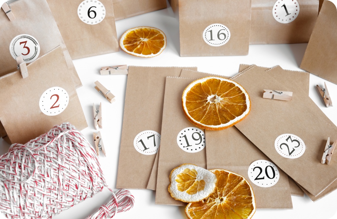 DIY-Adventskalender aus Papiertüten mit Zahlenstickern, Holzklammern, getrockneten Orangenscheiben und Bäckergarn auf weißem Hintergrund.