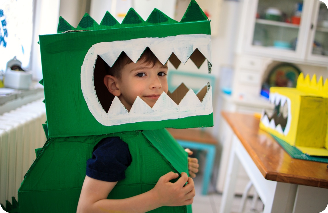 Lächelnder Junge trägt ein selbstgebasteltes Dinosaurier-Kostüm aus grün bemalten Kartons mit weißen Zähnen. Kreatives DIY-Projekt.