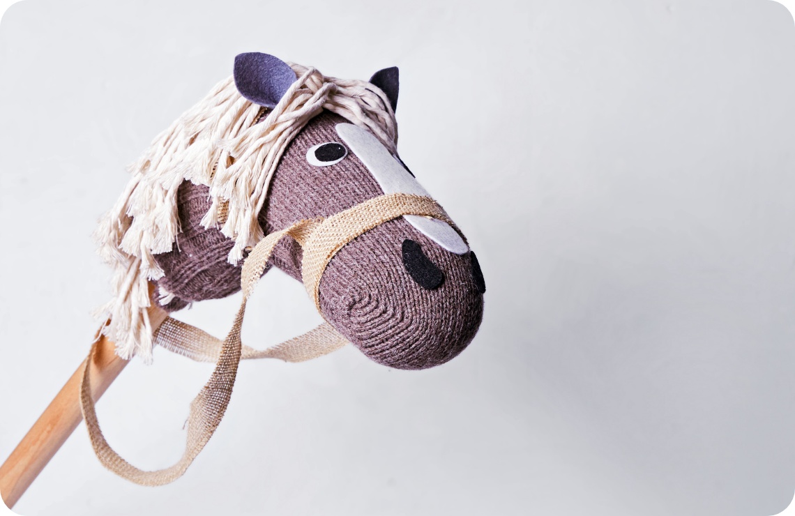 Selbstgemachtes Steckenpferd mit gestricktem Kopf, Jutegeschirr und heller Wollmähne vor neutralem Hintergrund. Kreatives DIY-Spielzeug.
