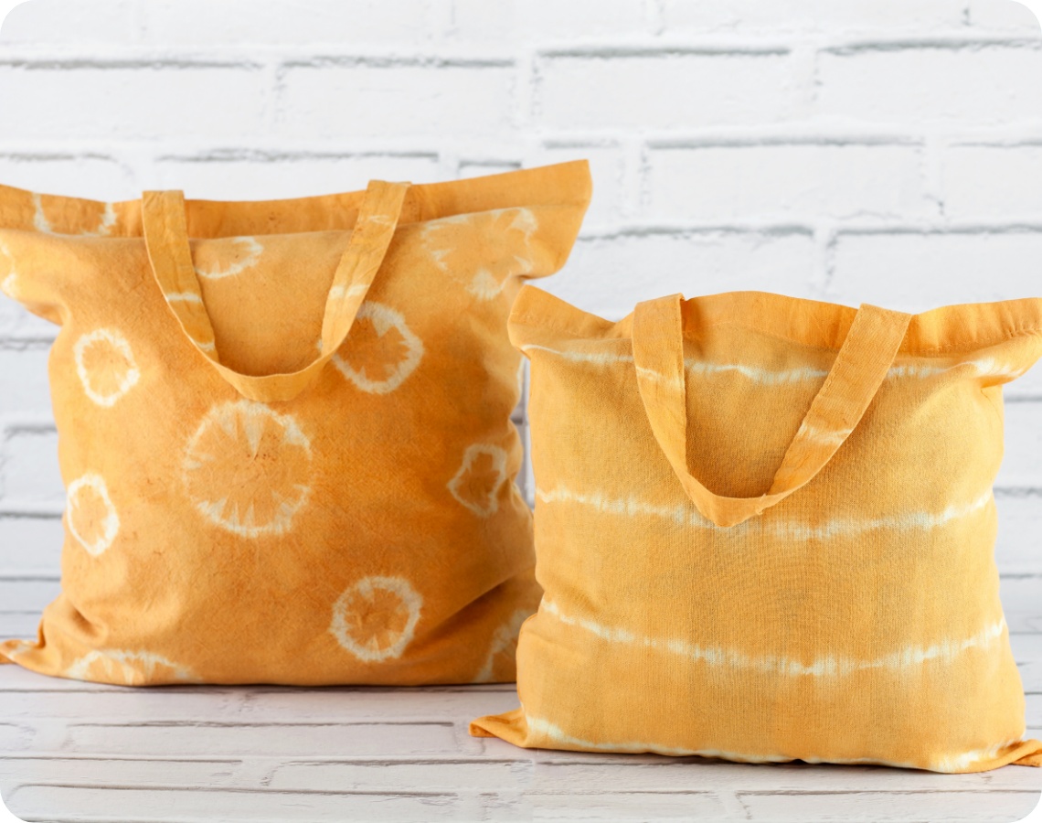 Zwei handgefärbte Stofftaschen in Gelb mit Batikmustern vor weißer Ziegelwand. Nachhaltige DIY-Taschen mit individuellem Design.