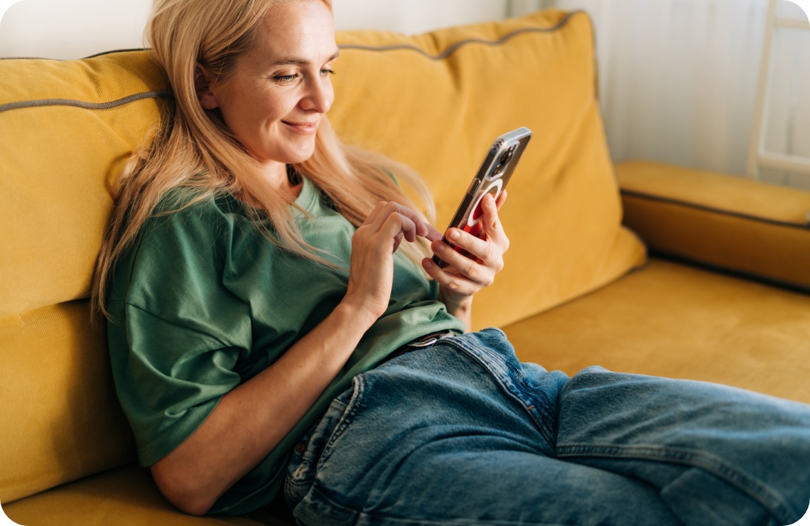 Frau in grünem T-Shirt liegt entspannt auf gelbem Sofa und benutzt ihr Smartphone.