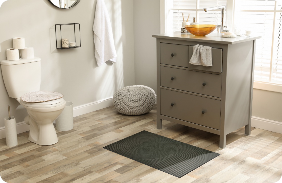 Modernes Badezimmer mit grauem Waschtisch, dunkelgrauer ALDI Fußmatte, Toilette und rundem Hocker.