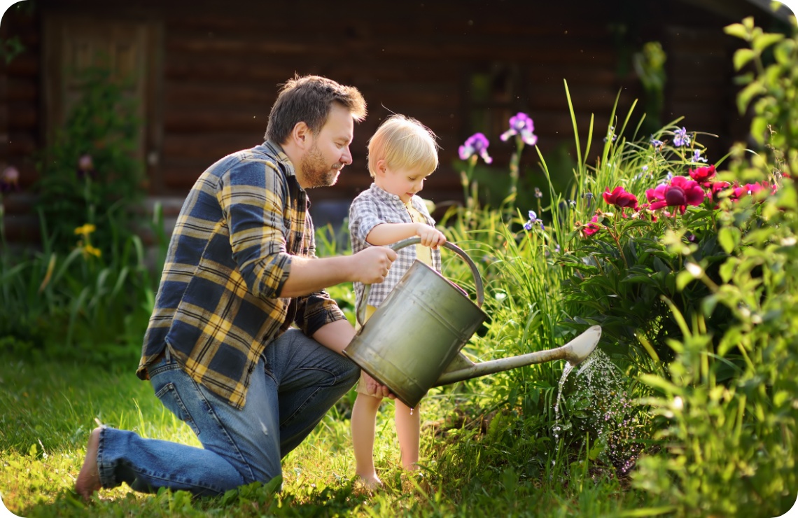 Vater und Kind gießen mit Metallkanne bunte Blumen im sonnigen Garten vor Holzhütte.
