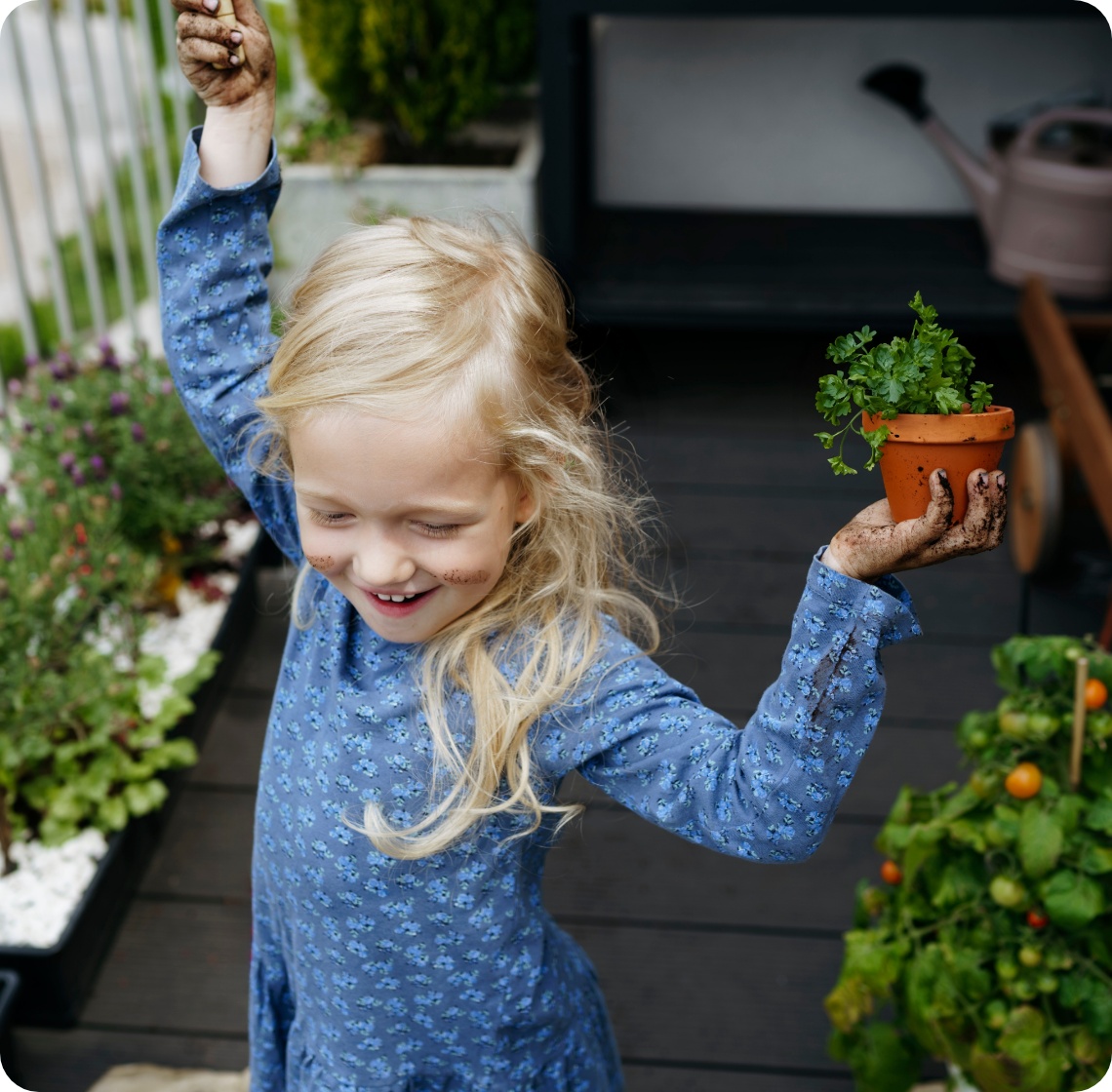 Fröhliches Mädchen mit Erde an den Händen hält Kräutertopf auf Balkon mit Gemüse- und Blumenpflanzen.
