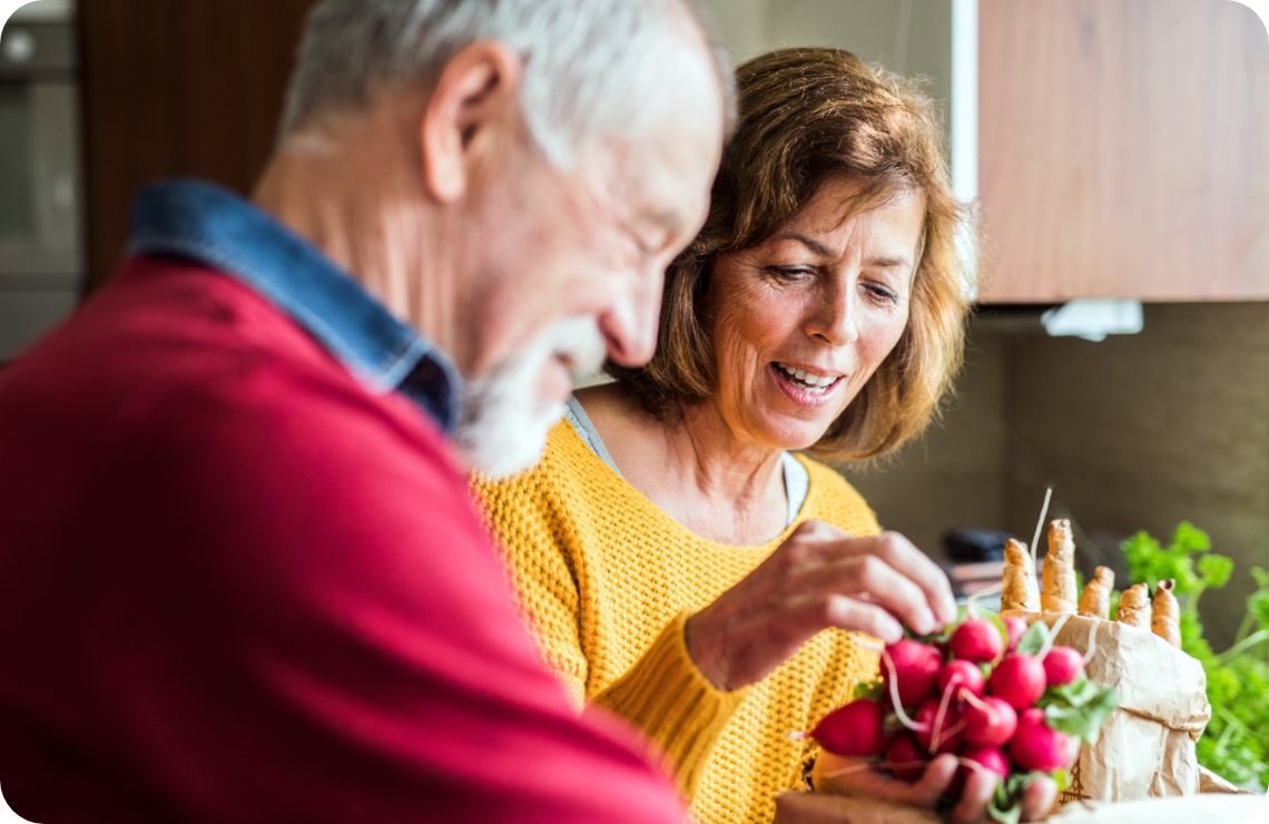 Älteres Paar in der Küche packt frische Radieschen und Karotten aus einer Papiertüte aus.