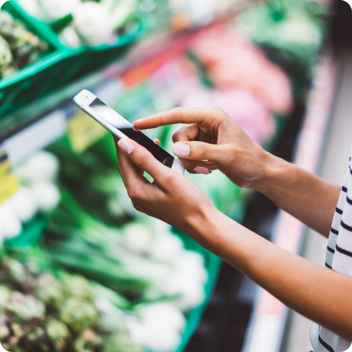Person im Supermarkt tippt auf ein Smartphone, während sie vor einem Regal mit frischem Gemüse steht.