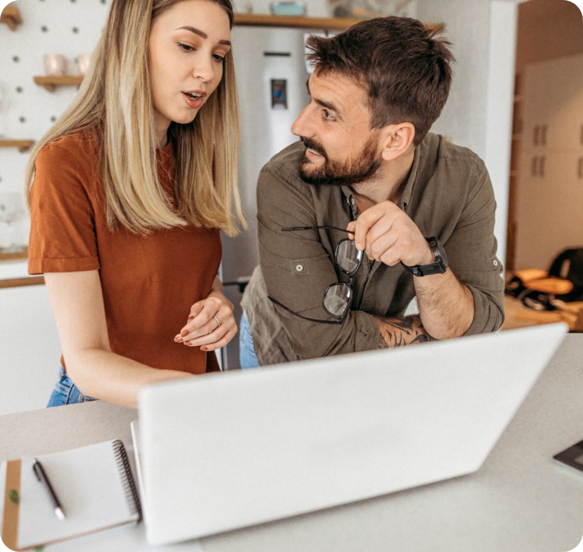 Paar bespricht etwas am Laptop in einer modernen Küche. Notizbuch und Tablet liegen auf dem Tisch.