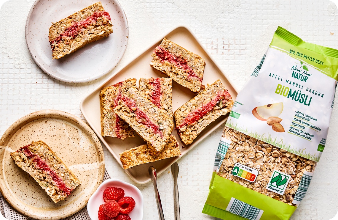 Bio-Müsliriegel mit Himbeerfüllung auf Tellern, daneben eine Packung Apfel-Mandel-Urkorn-Biomüsli und frische Himbeeren.