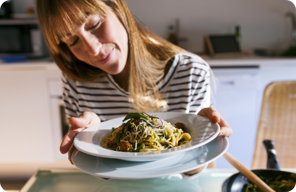 Frau präsentiert stolz einen Teller Spaghetti mit Gemüse und Käse in einer hellen Küche.