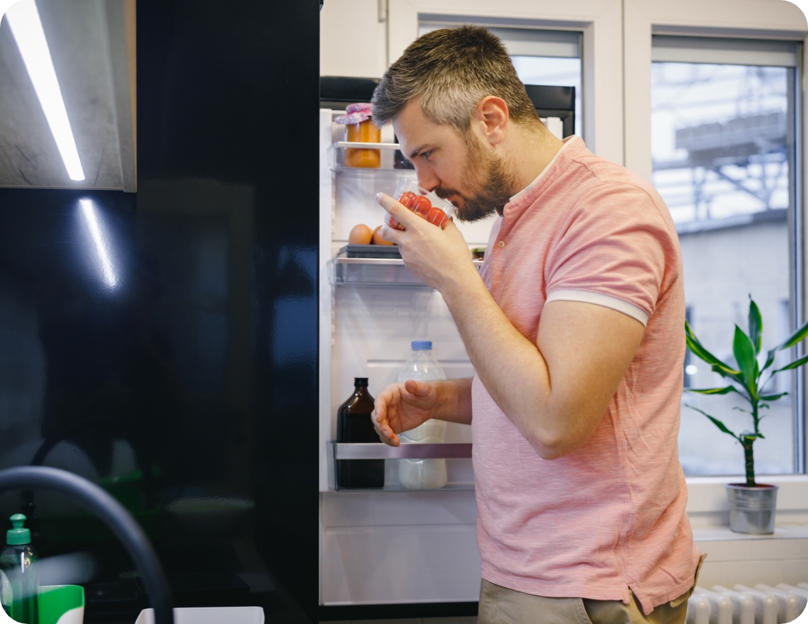 Mann in rosa Polohemd riecht an Tomaten vor offenem Kühlschrank.