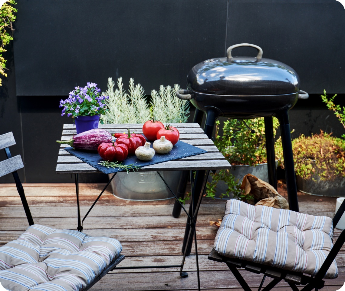 Gemüseauswahl mit Tomaten, Aubergine und Pilzen auf Holztisch vor Kugelgrill auf bepflanzter Terrasse.