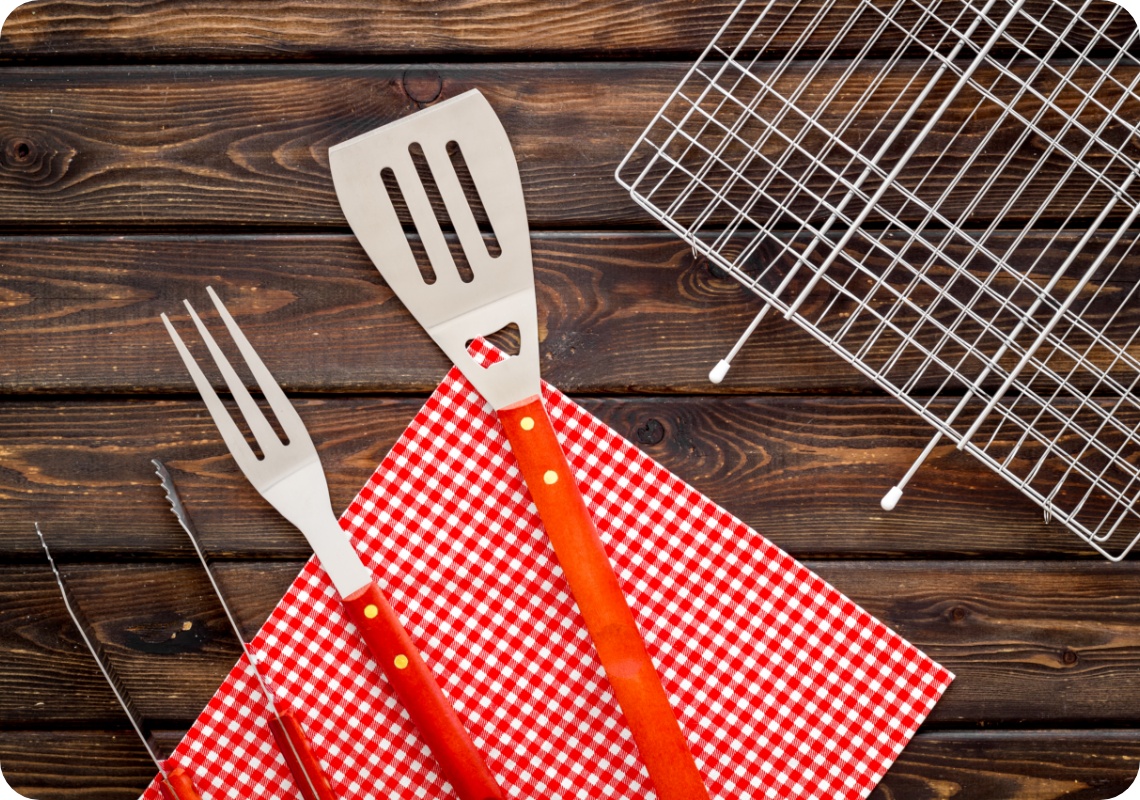 Grillbesteck mit Gabel, Zange und Wender auf rot-weißem Tuch, daneben Grillrost auf Holzuntergrund.