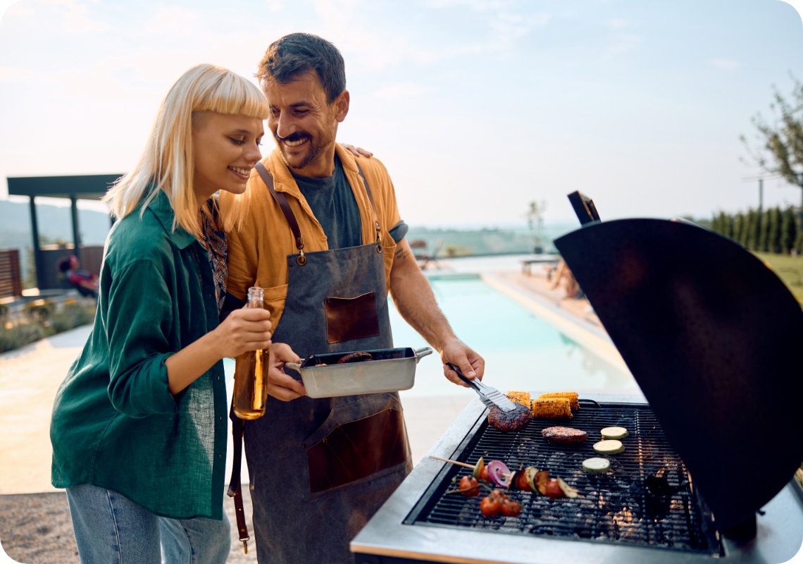 Lächelndes Paar beim Grillen am Pool, Mann in Schürze wendet Burger, Gemüse und Mais auf dem Grill.