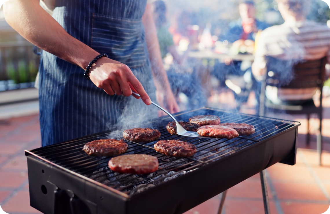 Person in gestreifter Schürze grillt mehrere Burger auf Holzkohlegrill, Rauch steigt auf, Gäste im Hintergrund.