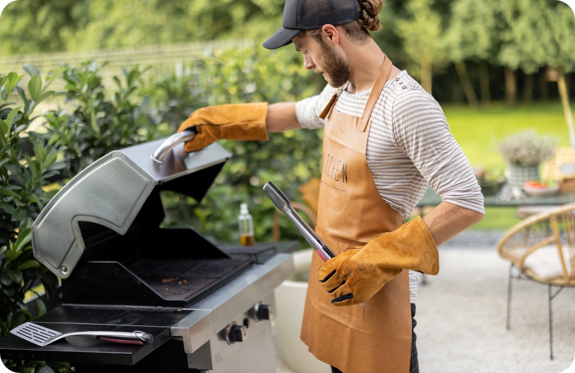 Mann mit Lederhandschuhen und Schürze öffnet Gasgrilldeckel im Garten, Grillzange in der Hand, bereit zum Grillen.