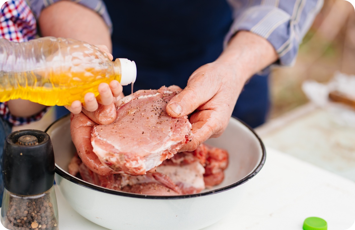 Rohes Schweinefleisch wird mit Öl beträufelt und gewürzt, Hände halten Fleisch über Schüssel, Pfeffermühle daneben.