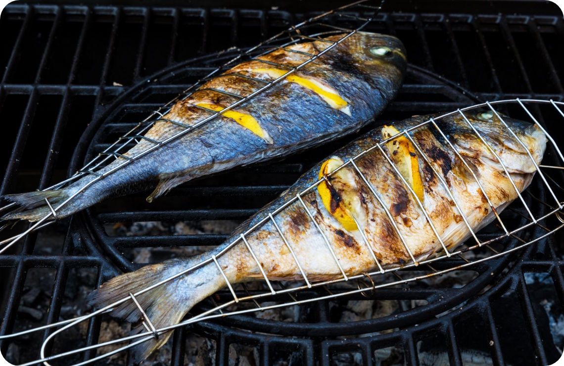 Zwei ganze Fische mit Zitronenscheiben in Grillgitter auf Holzkohlegrill, Haut goldbraun gegrillt.