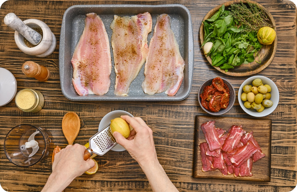 Roh marinierter Fisch in Auflaufform, umgeben von Kräutern, Oliven, getrockneten Tomaten und geriebener Zitrone auf Holztisch.