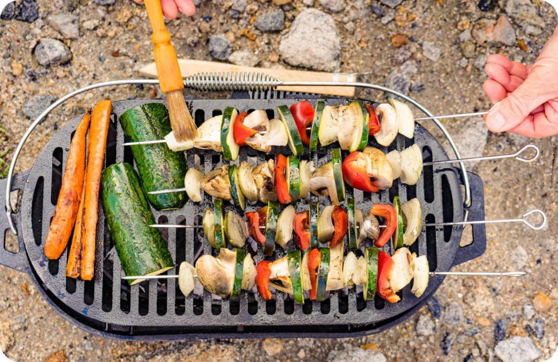 Gemüsespieße mit Paprika, Zucchini, Zwiebeln und Pilzen auf Grillrost, Möhren daneben, Marinade wird aufgetragen.