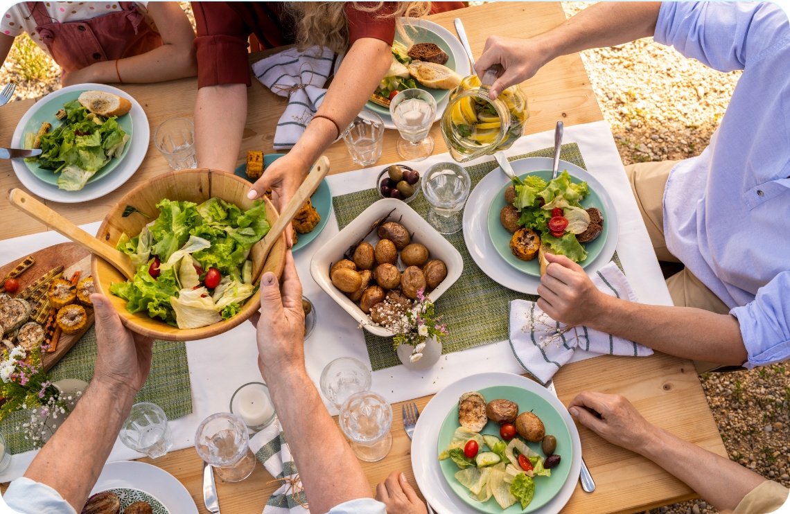 Sommerliche Tischszene mit Grillgemüse, Salat, Kartoffeln und Brot, mehrere Hände greifen zu, Wasser mit Zitrone wird eingeschenkt.