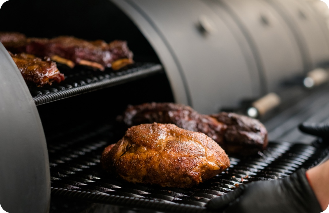 Großes Stück Fleisch mit Gewürzkruste im Smoker gegrillt, saftig gebräunt, weitere Fleischstücke auf oberem Rost.