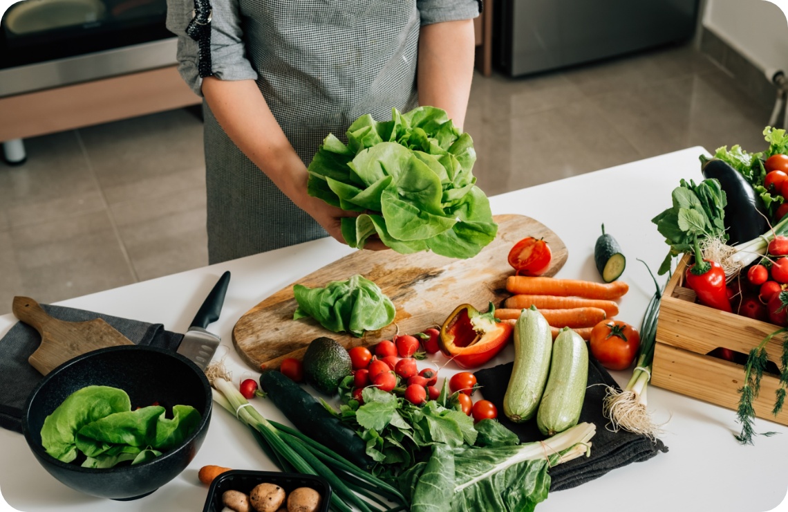 Person bereitet buntes Saisongemüse, wie Salat, Paprika, Gurken, Radieschen und Avocado auf einem Küchentisch vor.