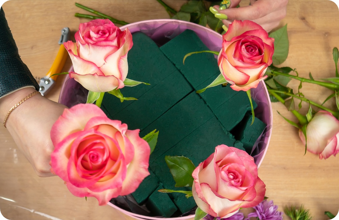 Person steckt vier rosa Rosen in einen Kreis mit grünem Steckschaum.