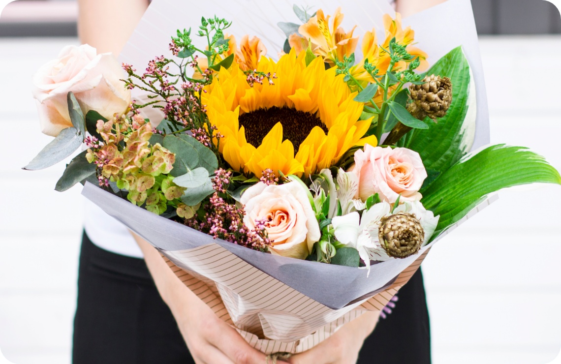 Person hält einen Blumenstrauß mit Rosen, Sonnenblumen und Grünzeug in der Hand.