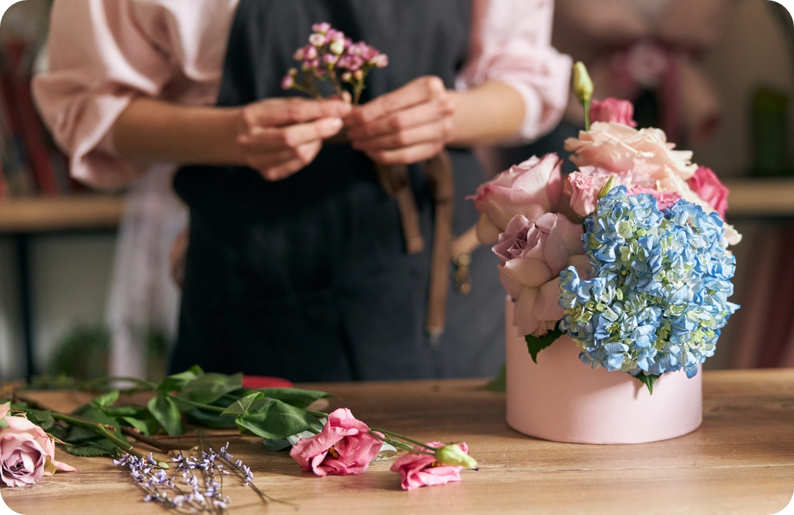 Floristin arrangiert zarte rosa Rosen und blaue Hortensien in pastellfarbener Hutschachtel auf Holztisch..