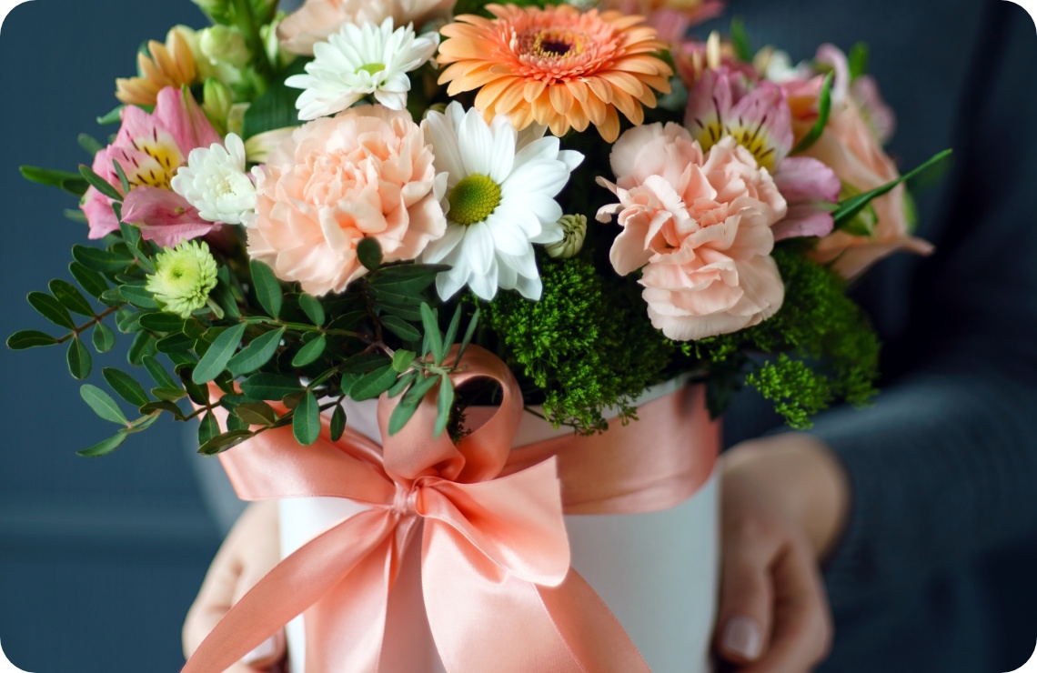 Runder Blumenstrauß mit pastellfarbenen Nelken, weißen Margeriten und rosa Schleife in Hutschachtel.