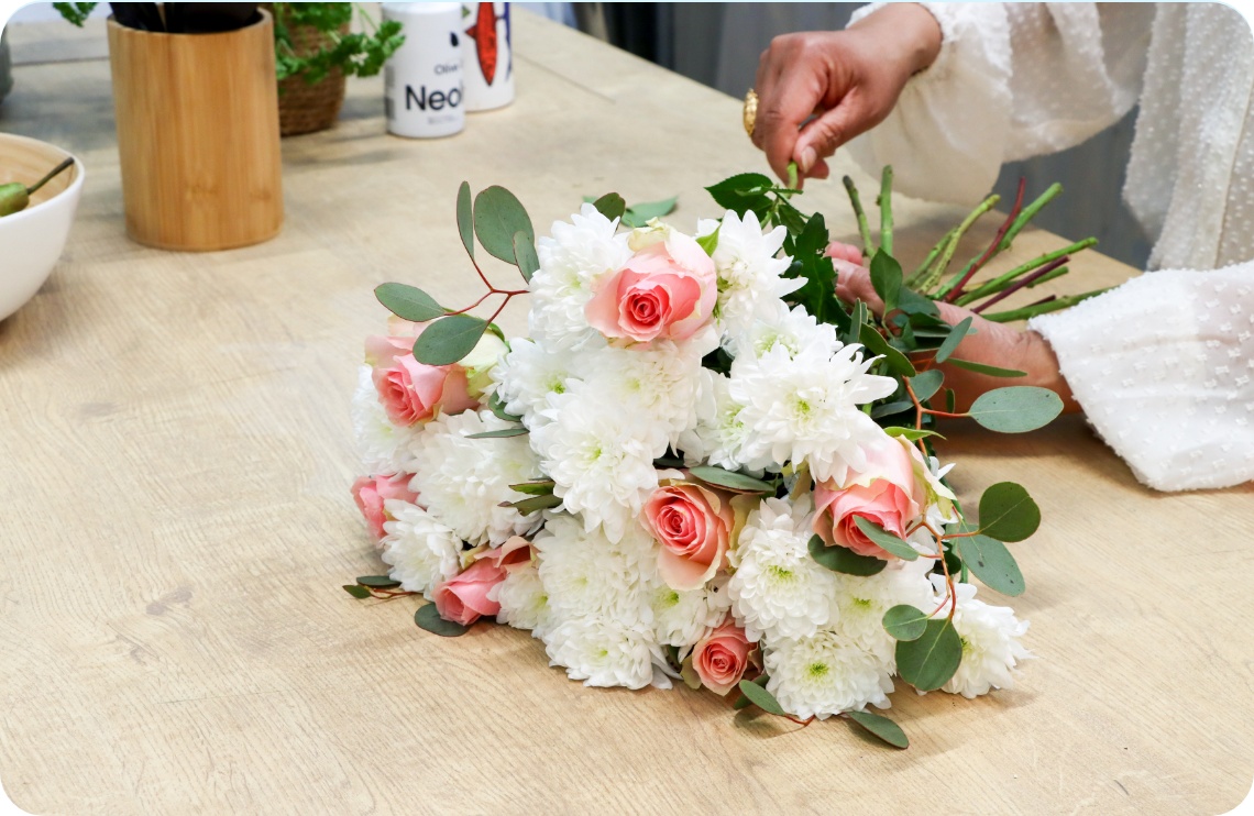Ein gebundener Blumenstrauß aus weißen Chrysanthemen, rosa Rosen und Eucalytptus liegt auf einer Tischplatte.