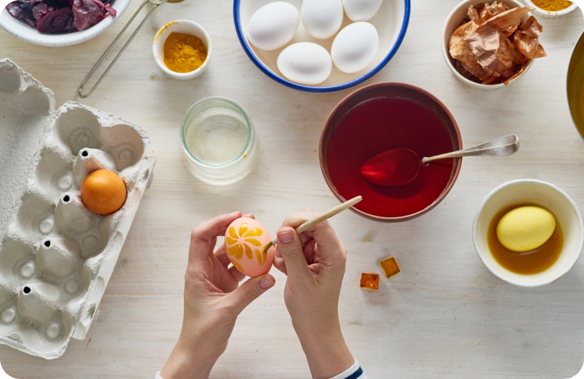 Person verziert ein Osterei mit einem floralen Muster. Auf einer weißen Arbeitsplatte stehen Kurkuma und Zwiebelschalen zum natürlichen Färben.