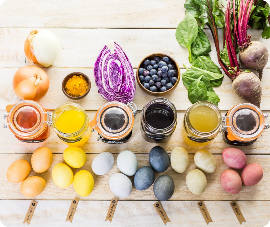 Natürliche Farbsude in Vorratsgläsern aus Zutaten wie Kurkuma, Rotkohl und Rote Bete mit gefärbten Eiern auf einem Holztisch arrangiert.