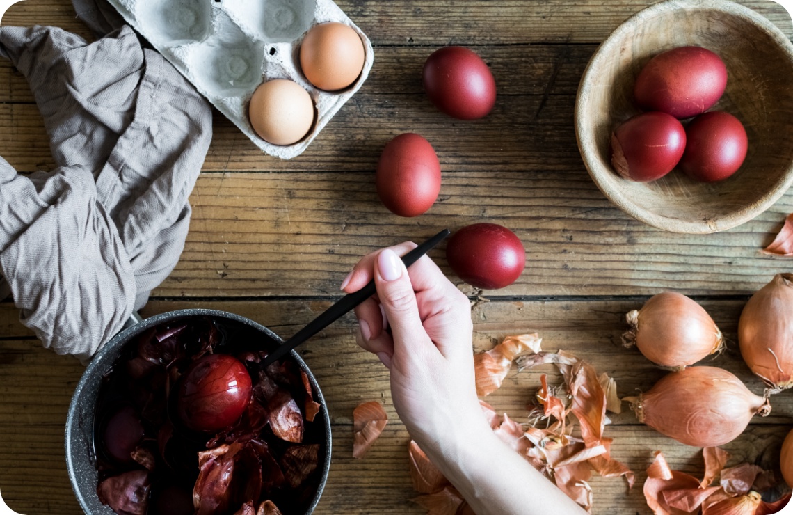 Person hebt ein tiefrot gefärbtes Osterei aus einem Topf mit Zwiebelschalen, umgeben von Eiern und Schalen auf Holztisch.