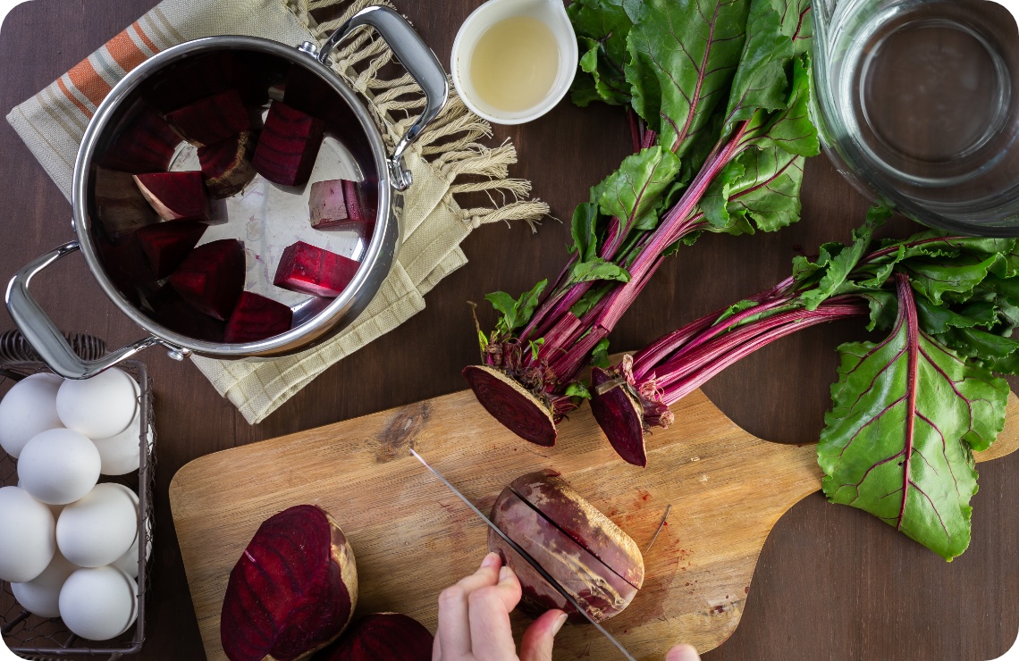 Person schneidet frische Rote Bete auf einem Holzbrett und gibt sie in einen Topf mit Essig. Auf dem Tisch stehen weiße Eier.