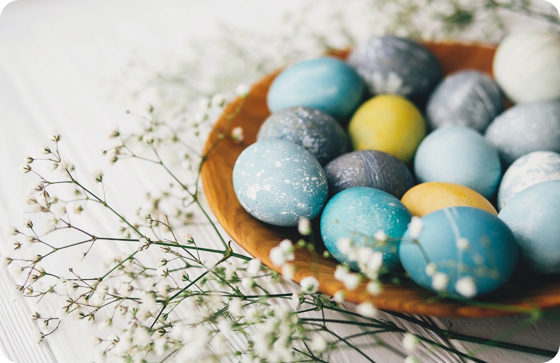 Bunter Ostereier-Mix in Blau, Gelb und Grau auf einem Holzteller, dekoriert mit zarten weißen Blüten.