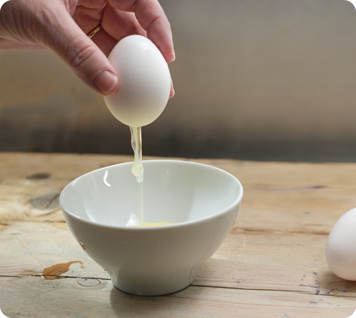 Eine Person bricht ein Ei über einer weißen Schüssel auf, während das Eiweiß hinabfließt. Zwei ganze Eier liegen daneben.