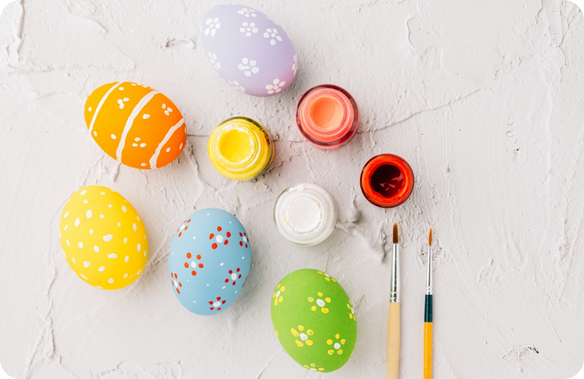 Bunt bemalte Ostereier mit Blumenmustern, Pinsel und Farbtöpfe auf strukturiertem weißen Hintergrund.