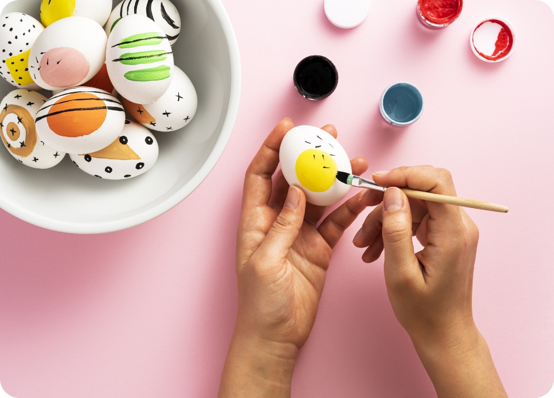 Person bemalt ein Osterei mit gelbem Kreis und schwarzen Strichen, daneben eine Schüssel mit modern verzierten Eiern.