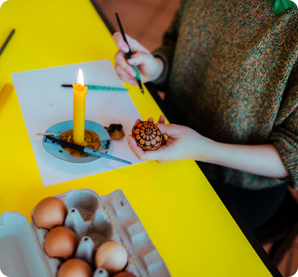 Person gestaltet ein Osterei mit Wachsbatik-Technik, gelbe angezündete Kerze steht auf dem Tisch.